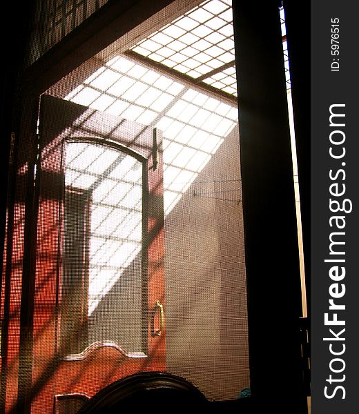 A photo taken of a wood door through a metal net with both sun n shadow. A photo taken of a wood door through a metal net with both sun n shadow