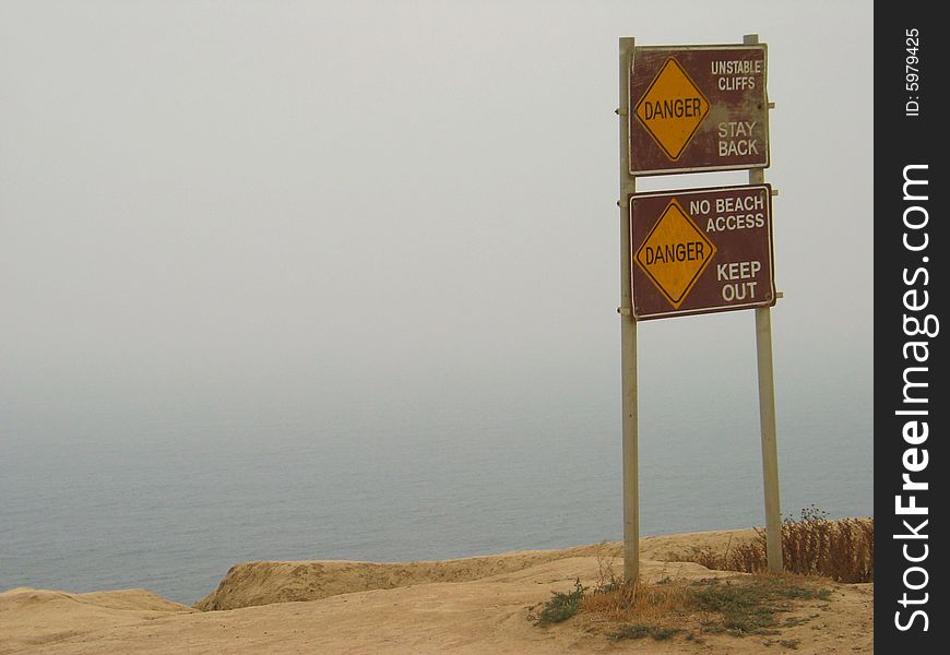 A sign on the edge of a cliff overlooking the ocean. The sign warns: Danger, Stay Back, Unstable Cliffs, No Beach Access, Keep Out. A sign on the edge of a cliff overlooking the ocean. The sign warns: Danger, Stay Back, Unstable Cliffs, No Beach Access, Keep Out.