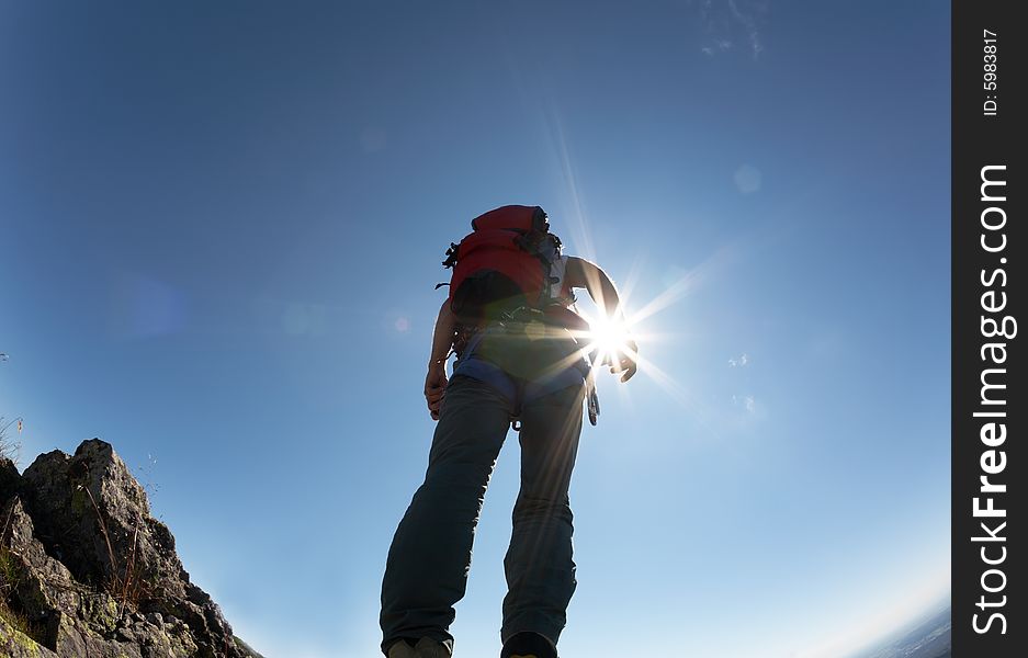 Climber, with climbing gears, standing on a stone at the top of his route, over a deep blue sky.