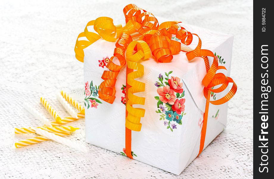 Beautiful gift box with vibrant orange and yellow ribbons sitting on a white lace tablecloth with birthday candles. Beautiful gift box with vibrant orange and yellow ribbons sitting on a white lace tablecloth with birthday candles.