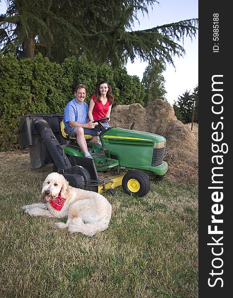 A man and woman sitting on a tractor smiling. Their dog is nearby on the grass. Vertically framed photograph. A man and woman sitting on a tractor smiling. Their dog is nearby on the grass. Vertically framed photograph