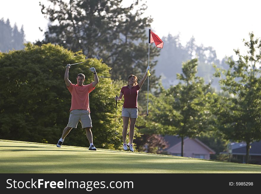 A young couple is playing on a golf course.  The man is holding his golf club above his head and the woman is holding the flag.  They are smiling at the camera.  Horizontally framed shot. A young couple is playing on a golf course.  The man is holding his golf club above his head and the woman is holding the flag.  They are smiling at the camera.  Horizontally framed shot.