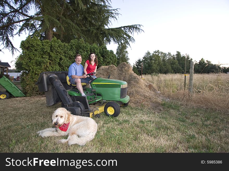 A man and woman sitting on a tractor smiling. Their dog is nearby on the grass. Horizontally framed photograph. A man and woman sitting on a tractor smiling. Their dog is nearby on the grass. Horizontally framed photograph
