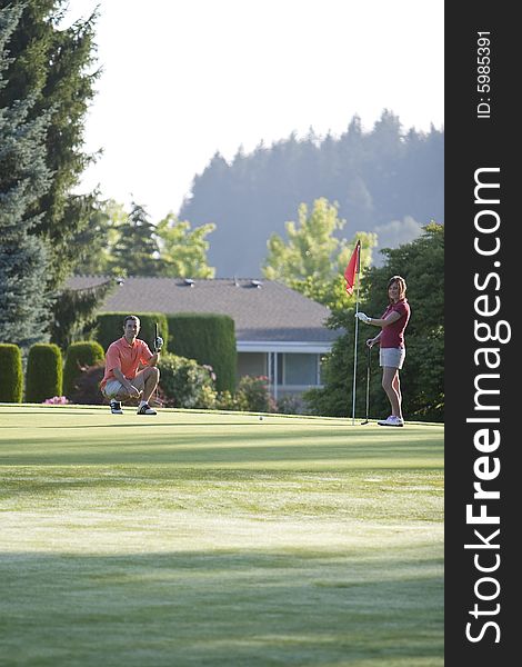 A young couple is setting up to play golf on the green of a golf course.  They are looking at the camera.  Vertically framed shot. A young couple is setting up to play golf on the green of a golf course.  They are looking at the camera.  Vertically framed shot.