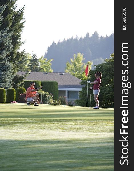 A young couple is setting up to play golf on the green of a golf course.  They are looking away from the camera.  Vertically framed shot. A young couple is setting up to play golf on the green of a golf course.  They are looking away from the camera.  Vertically framed shot.