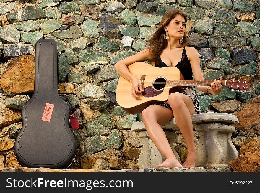 Pretty female singer playing guitar. A guitar case is resting against the rock wall.