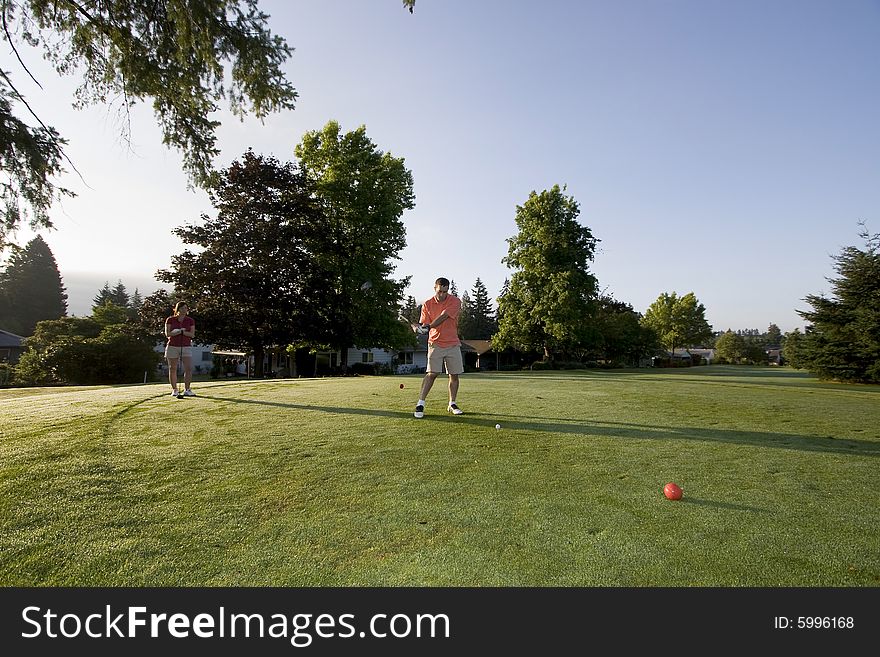 A young couple is standing on a golf course and playing golf.  The man is looking down at the ball he is swinging at and the woman is looking at the man.  Horizontally framed shot. A young couple is standing on a golf course and playing golf.  The man is looking down at the ball he is swinging at and the woman is looking at the man.  Horizontally framed shot.