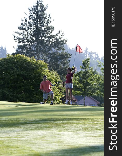 A young couple is playing on a golf course.  The man is holding his golf club and the woman is holding the flag.  They are looking away from the camera.  Vertically framed shot. A young couple is playing on a golf course.  The man is holding his golf club and the woman is holding the flag.  They are looking away from the camera.  Vertically framed shot.