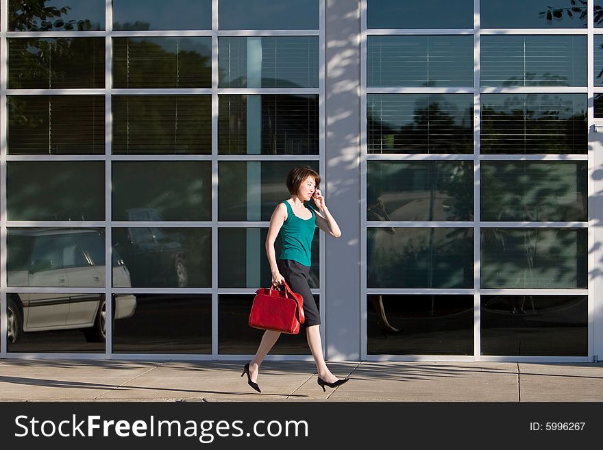 Yound Asian woman walks briskly while talking on the cell phone.  She is carrying a red handbag. Horizontally framed photo. Yound Asian woman walks briskly while talking on the cell phone.  She is carrying a red handbag. Horizontally framed photo.