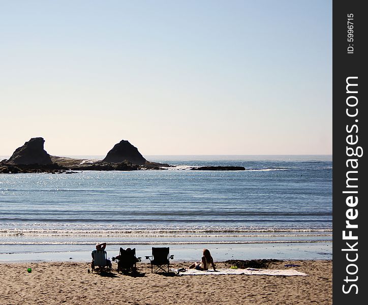 A view of rocky coastline from the beach, with people relaxing and looking off toward the horizon. A view of rocky coastline from the beach, with people relaxing and looking off toward the horizon.