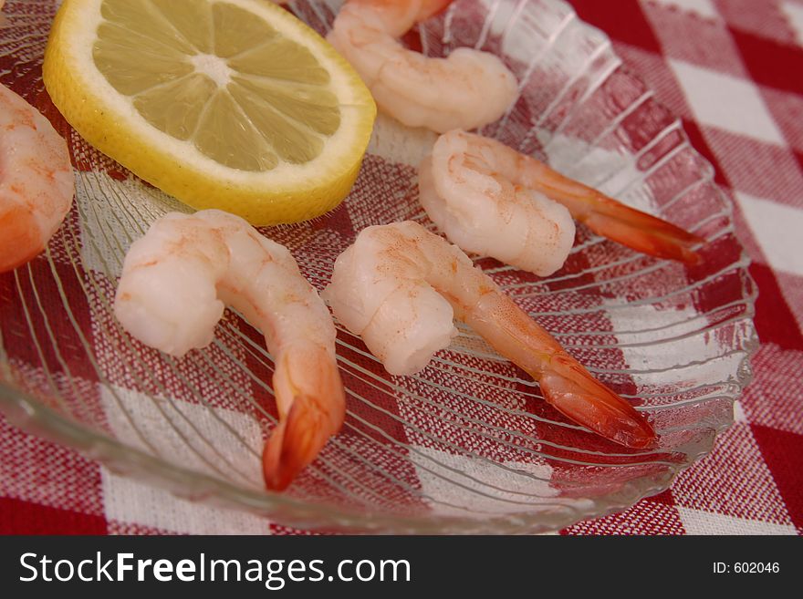 Boiled shrimp and lemon slice displayed on clear glass plate on red and white tablecloth.