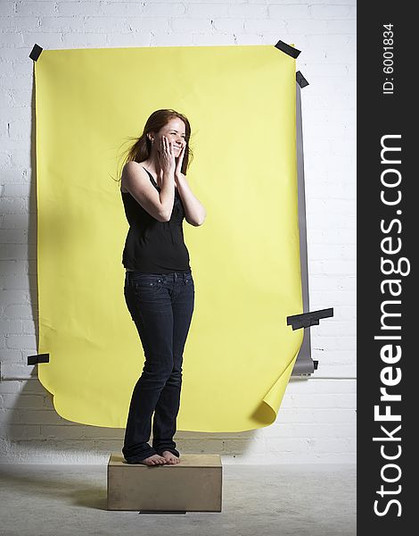 Young red headed woman standing on wooden box in front of yellow paper background laughs so much her cheeks hurt. Young red headed woman standing on wooden box in front of yellow paper background laughs so much her cheeks hurt.