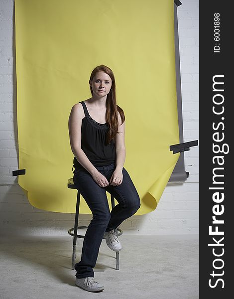 Young red headed woman sitting on a metal stool in front of a yellow background. Young red headed woman sitting on a metal stool in front of a yellow background.