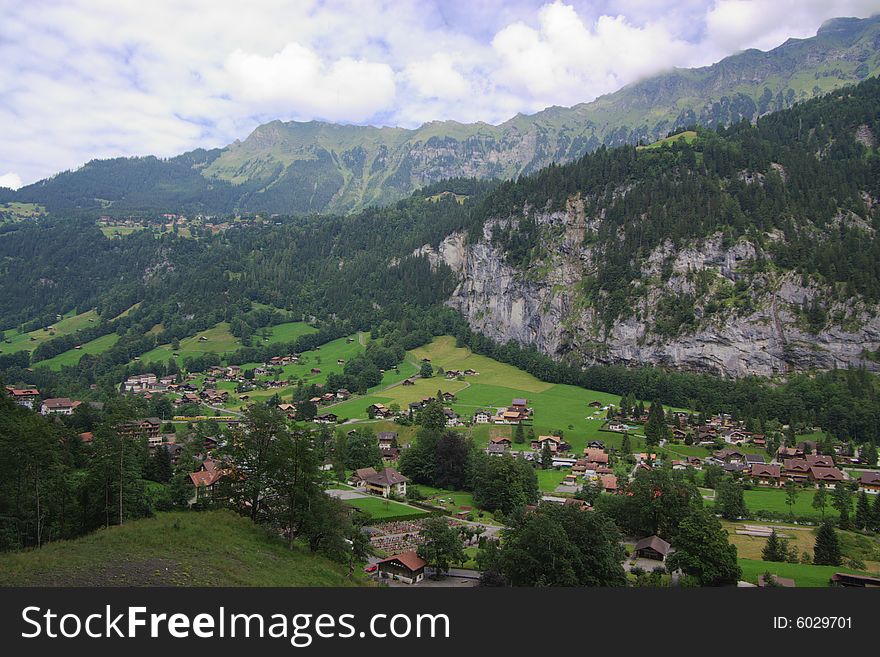 A panoramic view on green mountains under clouds with green slopes, small detached houses. Alpes, Switzerland, Europe. A panoramic view on green mountains under clouds with green slopes, small detached houses. Alpes, Switzerland, Europe.