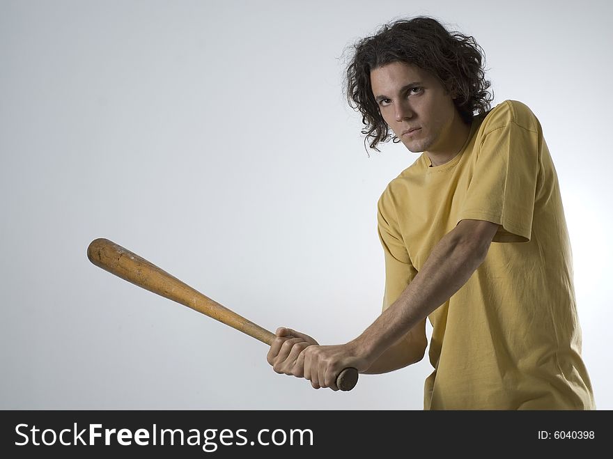 A young man in a yellow shirt, looking at the camera with a determined expression on his face, swinging a wooden bat. Horizontally framed shot. A young man in a yellow shirt, looking at the camera with a determined expression on his face, swinging a wooden bat. Horizontally framed shot.