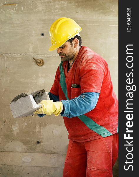 Construction worker holds cinder block while layering cement. He is wearing an orange suit and a yellow hard hat. Vertically framed photo. Construction worker holds cinder block while layering cement. He is wearing an orange suit and a yellow hard hat. Vertically framed photo.
