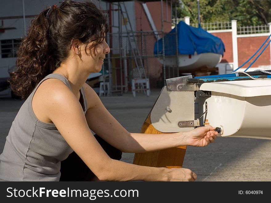 A young woman is kneeling and fixing something on her sailboat.  She is smiling lightly and looking away from the camera.  Horizontally framed shot. A young woman is kneeling and fixing something on her sailboat.  She is smiling lightly and looking away from the camera.  Horizontally framed shot.