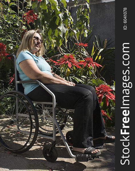 Woman in blue shirt reclines in wheelchair next to flowery bush. She is wearing sunglasses and looking ahead. Vertically framed photo. Woman in blue shirt reclines in wheelchair next to flowery bush. She is wearing sunglasses and looking ahead. Vertically framed photo.