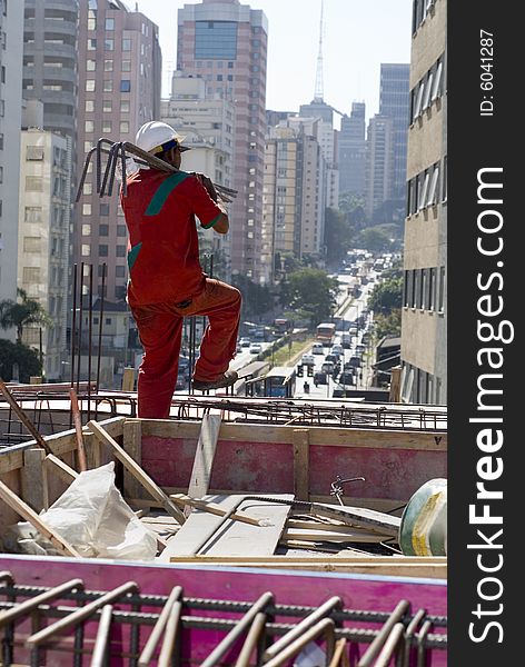 Construction worker in orange suit carries rebar over his shoulder while working on top of building. Vertically framed photo. Construction worker in orange suit carries rebar over his shoulder while working on top of building. Vertically framed photo.