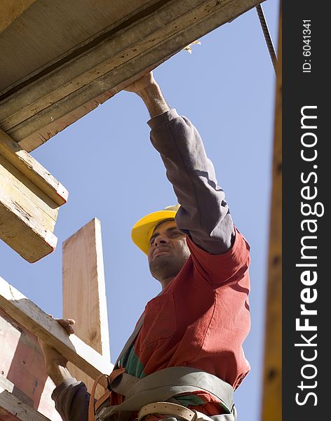 Construction worker carries board up ladder. He is wearing an orange suit and a yellow hard hat. Vertically framed photo. Construction worker carries board up ladder. He is wearing an orange suit and a yellow hard hat. Vertically framed photo.