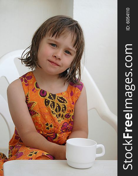 Portrait of the little girl In an orange dress sitting at a table and a white cup. Portrait of the little girl In an orange dress sitting at a table and a white cup