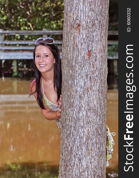 A young lady peeks around a tree with a lake and foot bridge in the background. A young lady peeks around a tree with a lake and foot bridge in the background.