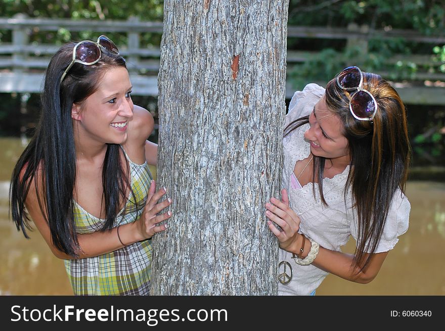 Two young ladies peeks around a tree with a lake and foot bridge in the background. Two young ladies peeks around a tree with a lake and foot bridge in the background.