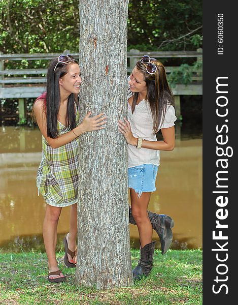 Two young ladies peeks around a tree with a lake and foot bridge in the background. Two young ladies peeks around a tree with a lake and foot bridge in the background.