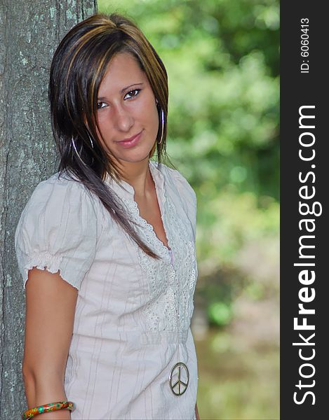 A young lady poses by a tree looking straight at the camera, head slightly tilted. A young lady poses by a tree looking straight at the camera, head slightly tilted