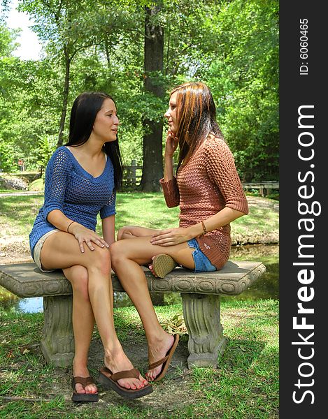 Two young ladies sit on a park bench, smiling and talking. Two young ladies sit on a park bench, smiling and talking.