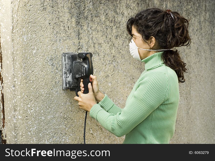 Woman wearing a paint mask uses an electric sander to sand a wall. Horizontally framed photo. Woman wearing a paint mask uses an electric sander to sand a wall. Horizontally framed photo.