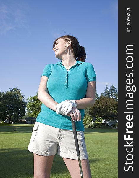 A young woman is standing on a golf course holding a club.  She is smiling, laughing, and looking away from the camera.  Vertically framed shot. A young woman is standing on a golf course holding a club.  She is smiling, laughing, and looking away from the camera.  Vertically framed shot.