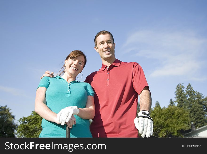 A young couple is standing on a golf course.  The man has his arm around the woman and they are smiling at the camera.  Horizontally framed shot. A young couple is standing on a golf course.  The man has his arm around the woman and they are smiling at the camera.  Horizontally framed shot.