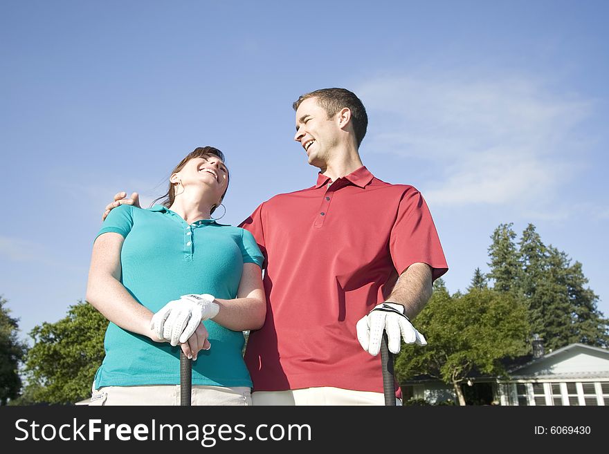 A young couple is standing on a golf course.  The man has his arm around the woman and they are laughing and looking away from the camera.  Horizontally framed shot. A young couple is standing on a golf course.  The man has his arm around the woman and they are laughing and looking away from the camera.  Horizontally framed shot.