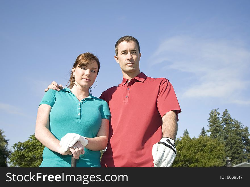 A young couple is standing on a golf course.  The man has his arm around the woman and they are looking sternly at the camera.  Horizontally framed shot. A young couple is standing on a golf course.  The man has his arm around the woman and they are looking sternly at the camera.  Horizontally framed shot.