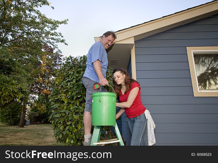 Man and woman standing on a ladder near a blue house. She is looking in a bucket and grimacing. Horizontally framed shot. Man and woman standing on a ladder near a blue house. She is looking in a bucket and grimacing. Horizontally framed shot.