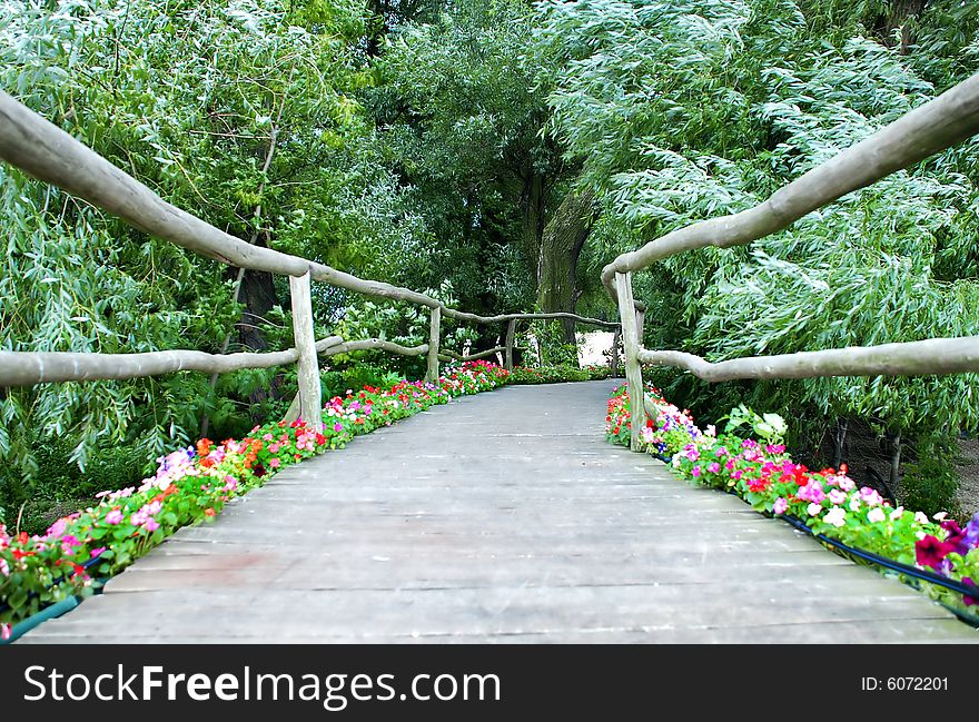 Wooden bridge with rails and flowers between green trees. Wooden bridge with rails and flowers between green trees