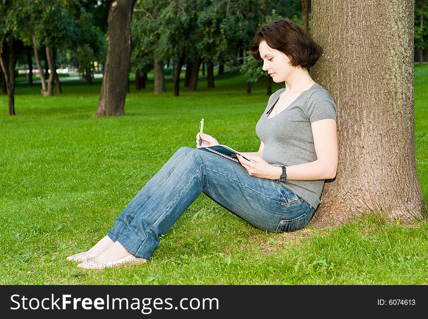 Girl sits under a tree in park and writes to notebook. Girl sits under a tree in park and writes to notebook
