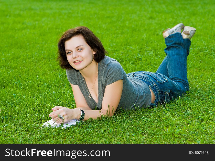 Smiling girl lays on a green grass with a notebook and a pen. Smiling girl lays on a green grass with a notebook and a pen