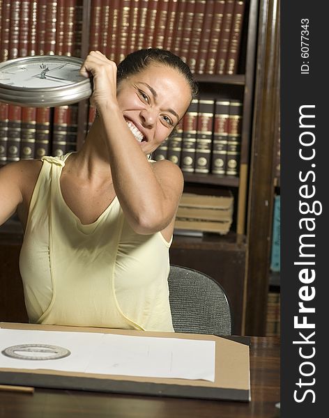 Woman being silly pretending to be mad while she holds a clock. There are books and paperwork surrounding her. Vertically framed photograph. Woman being silly pretending to be mad while she holds a clock. There are books and paperwork surrounding her. Vertically framed photograph.