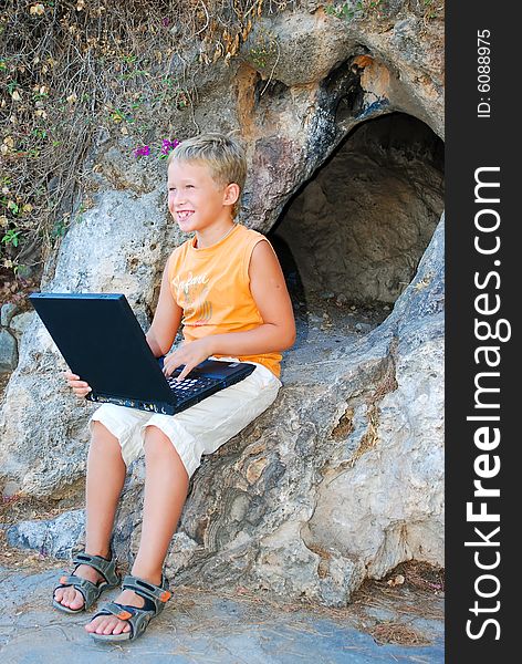 Cheerful boy plays on laptop near a cave