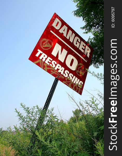 A shaded, red, metal sign reading Danger No Trespassing with graffiti markings on it, leaning out of the ground with green weeds, plants, and tree leaves behind it, against a clear blue sky