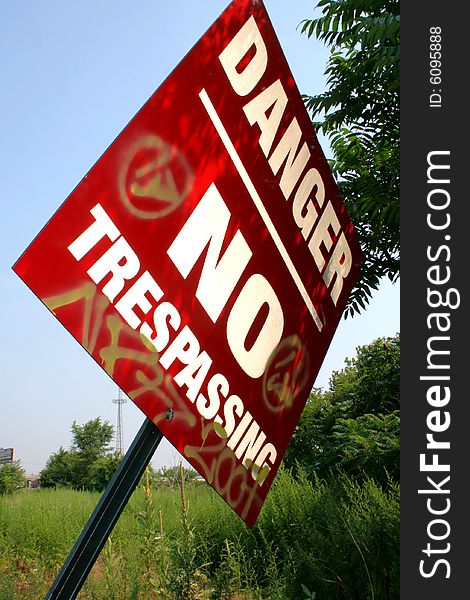 A shaded, red, metal sign reading Danger No Trespassing with graffiti markings on it, leaning out of the ground with a field of green weeds, plants, and tree leaves behind it, against a clear blue sky