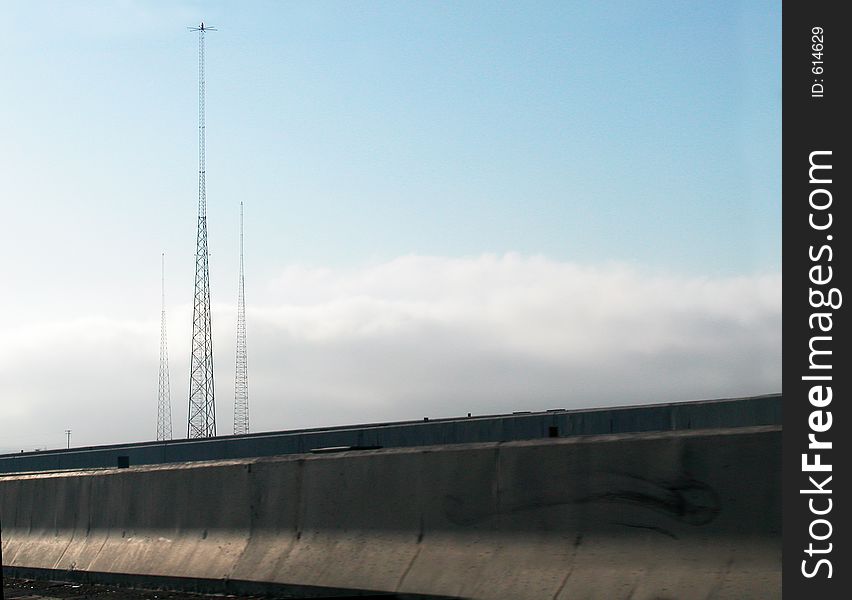 Viewed from a freeway tall radio towers are visible above concrete barriers and clouds. Viewed from a freeway tall radio towers are visible above concrete barriers and clouds