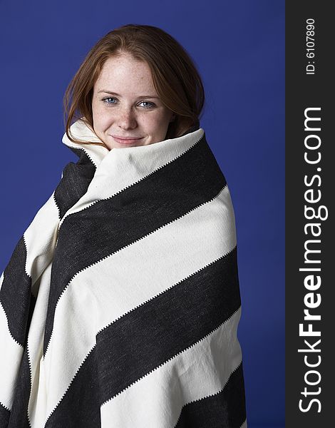 Young beautiful red headed woman with freckels and blue eyes wraps herself in a striped blanket.  She is against a deep blue background. Young beautiful red headed woman with freckels and blue eyes wraps herself in a striped blanket.  She is against a deep blue background.