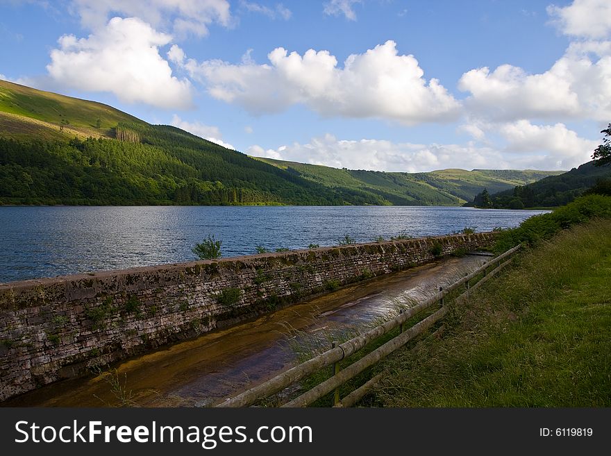 Lake with crease used to pour off water lit by sun and surrounded by green hills. Lake with crease used to pour off water lit by sun and surrounded by green hills