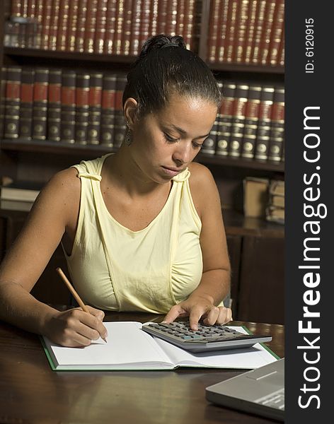 Woman sits at a table in the library. She is studying and has a calculator resting on her book.  Vertically framed photo. Woman sits at a table in the library. She is studying and has a calculator resting on her book.  Vertically framed photo.