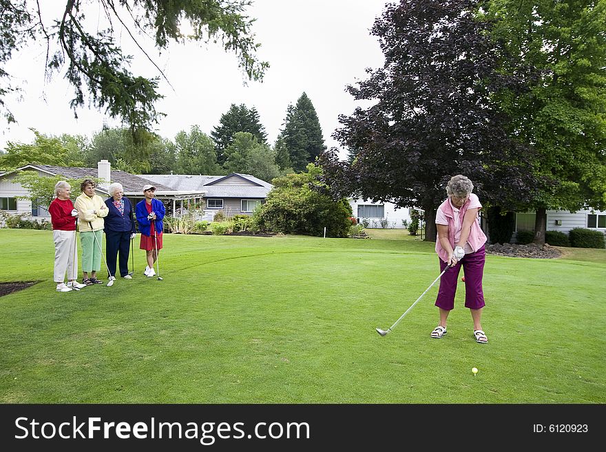Elderly woman teeing off on the golf course as her four friends watch. Horizontally framed photo.