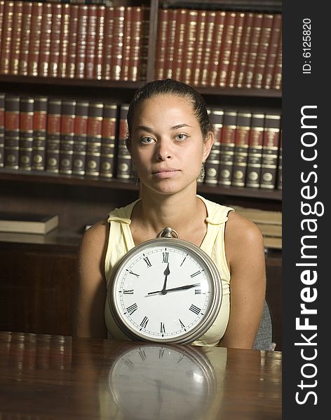 Woman sits at table in a library. She has a clock in front of her resting on table. Vertically framed photo. Woman sits at table in a library. She has a clock in front of her resting on table. Vertically framed photo.