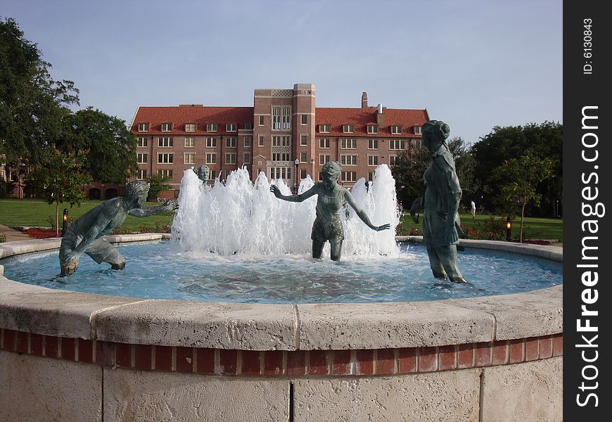 This is a water fountain that has bronze statues of people playing and having fun. There is a university dormitory in the back ground with blue skies.  This is a picture perfect college day. This is a water fountain that has bronze statues of people playing and having fun. There is a university dormitory in the back ground with blue skies.  This is a picture perfect college day.